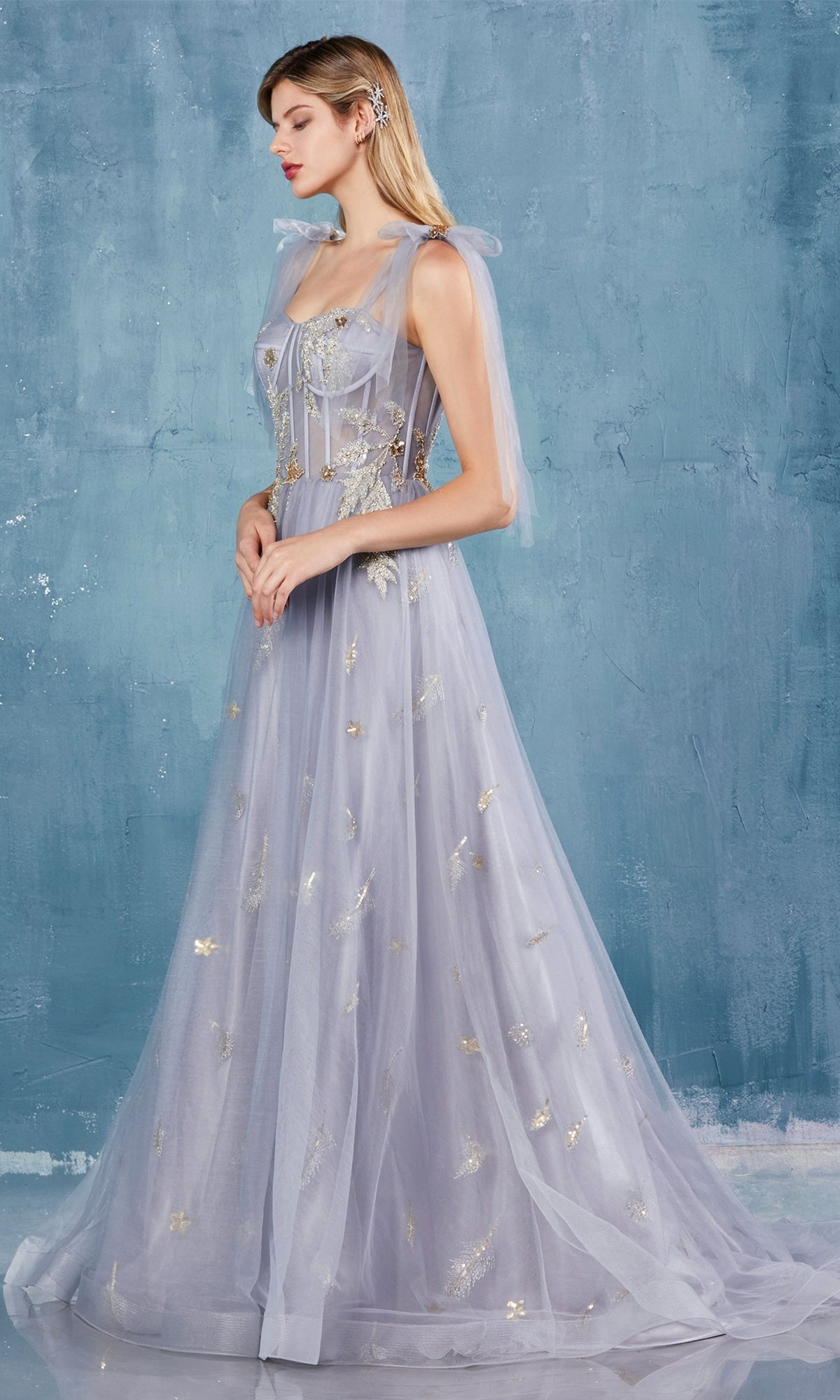 A woman stands in profile against a textured blue background, wearing the Shoulder-Bow Long Corset Prom Ball Gown A0824: a long light purple tulle dress with a corset bodice and gold floral embellishments.